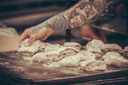 A baker with tattoos puts raw dough buns on the baking tray in the bakeryの写真素材
