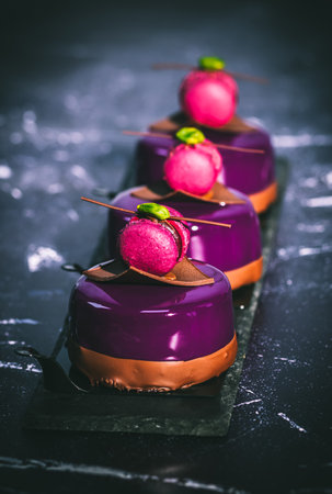 Three tartlets with purple creamy glaze, decorated with macaroon, chocolate and pistachio, on dark background, verticalの写真素材
