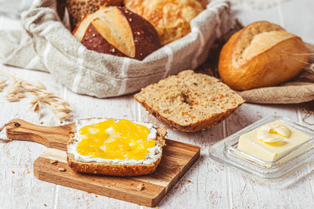 Half of a roll spread with cream cheese and lemon curd, variety of bread rolls in the background, scene of a breakfastの写真素材