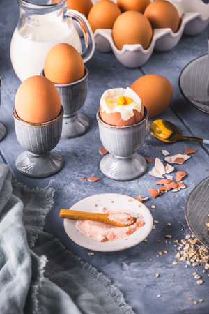 Boiled brown eggs in gray egg cups and other breakfast ingredients on gray background, preparation...