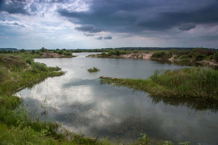 Thunderclouds over the lake. Reflection of the sky in the water. Wooden fishing boat on the shore of a pond.の写真素材