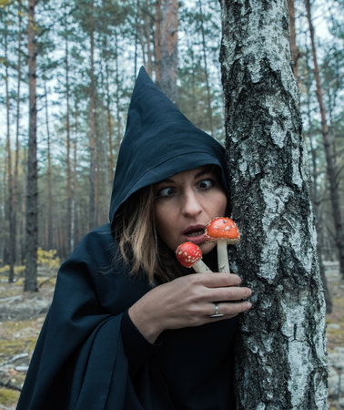A witch in a black hood and with red fly agarics conjures in the forest.