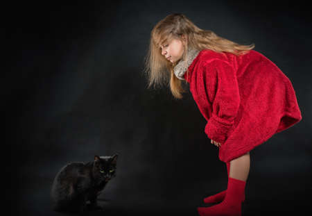 Teenage girl in a red fur coat in the studio on a black background with a black cat.の写真素材