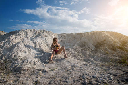 Summer heat. A girl in a bathing suit and sunglasses with a cocktail and pineapple in their hands drinking fresh juice from a straw against the background of white hills and cracked clay.の写真素材