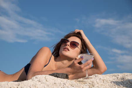 Summer heat. A girl in a bathing suit and sunglasses with a cocktail and pineapple in their hands drinking fresh juice from a straw against the background of white hills and cracked clay.の写真素材