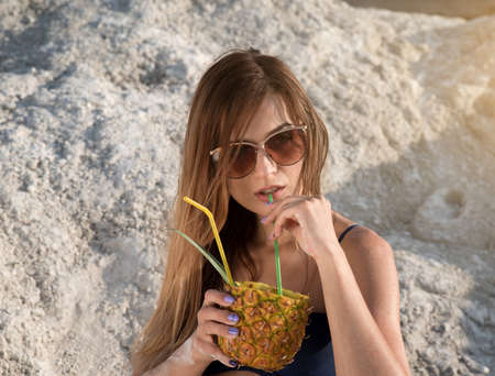 Summer heat. A girl in a bathing suit and sunglasses with a cocktail and pineapple in their hands drinking fresh juice from a straw against the background of white hills and cracked clay.の写真素材