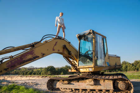 A girl in a white bodysuit in an open space among the sand near an old excavator.