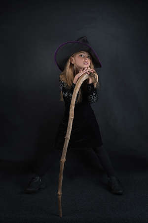A girl on a dark background in a witch costume and in a wide-brimmed hat with a staff for Halloween.の写真素材