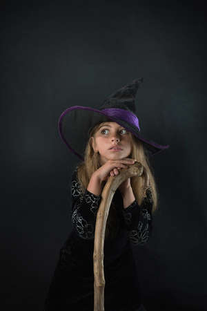 A girl on a dark background in a witch costume and in a wide-brimmed hat with a staff for Halloween.