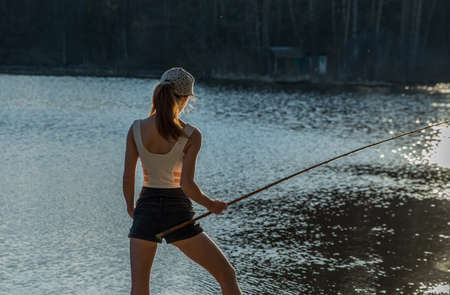 A girl stands on the shore of a forest lake and catches fish with a fishing rod.の写真素材