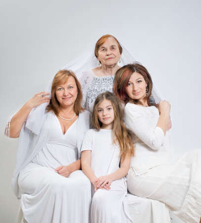 Soft group portrait of women of the same family of different generations in light clothes on a white...
