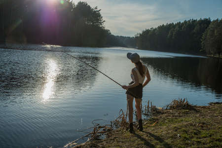 A girl stands on the shore of a forest lake and catches fish with a fishing rod.の写真素材