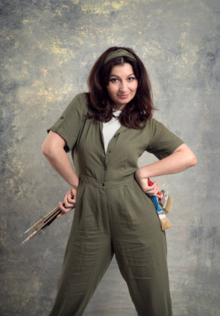 The artist is a woman in a green overalls and with brushes stained in paint on a studio background.の写真素材
