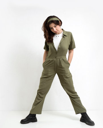 A woman on a light background in a green jumpsuit overalls stands in a pose with her legs apart and looks at the camera.の写真素材