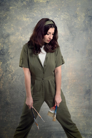 The artist is a woman in a green overalls and with brushes stained in paint on a studio background.の写真素材