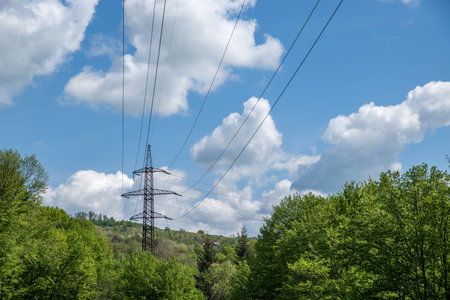 A high-voltage tower with wires among the trees against a cloudy blue sky.の写真素材