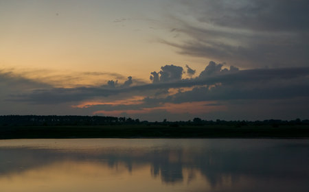 The evening dark sky with clouds illuminated by the setting sun over the dark shore of the reservoir.の写真素材