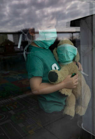 Coronavirus, Covid-19, Boy and his stuffed puppy in protection masks look through the window, quarantine at homeの写真素材