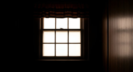 A square window with multiple panes is illuminated from the outside, casting a bright light into a dark room. Striped curtains hang above the window. To the right, a textured wooden wall is visible, with light reflecting off its vertical lines. The overall mood is one of quiet solitude.の写真素材