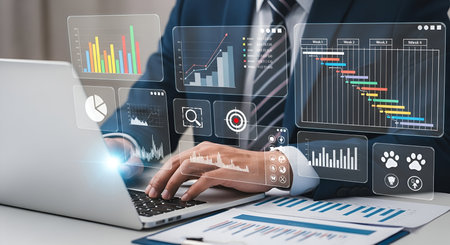A close-up shot of a person's hands working on a laptop, with a vibrant digital interface displaying various charts, graphs, and a Gantt chart. This visual emphasizes business analytics, data visualization, and performance tracking in a modern technological context.の写真素材