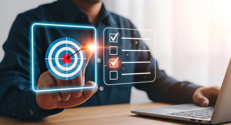 A man in a dark blue shirt is shown with his finger pointing towards a digital interface. The interface features a glowing blue outline of a target with an arrow hitting the bullseye, symbolizing achievement and focus. Next to the target is a checklist with several items, three of which are marked with red checkmarks, indicating completed tasks. A laptop is visible on the right.の写真素材