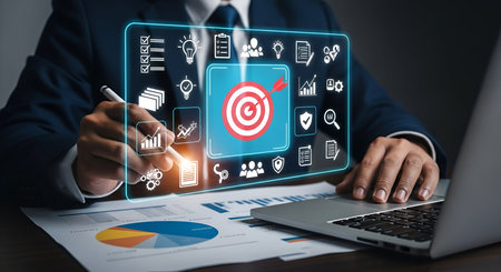 A man in a dark suit is using a stylus to interact with a glowing blue digital interface. The interface prominently displays a target with a dart, alongside icons for checklists, lightbulbs, documents, a shield, and team members. Charts and graphs are also visible. A laptop and papers with charts are on the desk.の写真素材