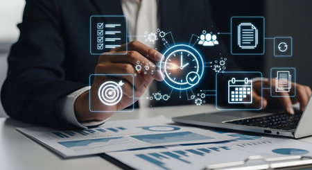 A person in a suit interacts with a digital interface showing icons for time management, including a clock, calendar, checklist, and gears. Charts and graphs are visible on documents below, and a laptop is on the right.の写真素材
