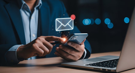 A person in a suit uses a smartphone, with a glowing envelope icon indicating a new email notification. The scene is set with a laptop in the foreground and blurred lights in the background, suggesting a focus on digital communication and connectivity.の写真素材