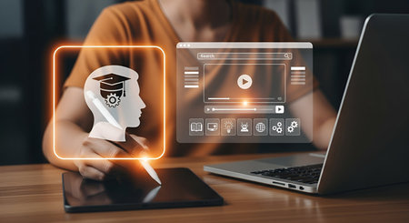 A person is using a tablet and a stylus to interact with a digital interface displaying an icon of a head with a graduation cap and gears, symbolizing learning and knowledge. Adjacent to it is a virtual window showing a video player and various educational icons. A laptop is also present.の写真素材
