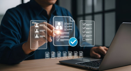 A person is shown interacting with a digital interface that displays lists of profiles and documents. One central document shows a profile with checkboxes and a large blue checkmark, indicating completion or approval. The person is using a stylus to interact with the screen, with a laptop in the background.の写真素材