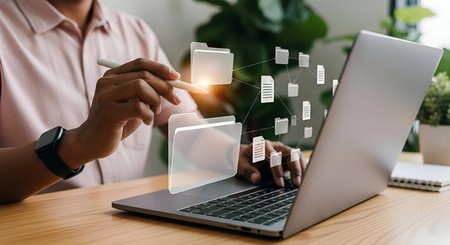 A person sits at a wooden desk, interacting with a laptop. Their hand holds a stylus, pointing at a holographic interface displaying a network of digital folders and documents. The laptop screen shows a keyboard and a portion of the screen. The scene suggests digital file management, data organization, and technological interaction.の写真素材