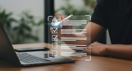 A person's hand holds a stylus and is in the process of marking a checkmark on a digital checklist displayed on a transparent interface. The checklist has several items, with three already marked as complete. A laptop and a smartphone are visible on the wooden desk in the foreground, suggesting a work or organizational context.の写真素材