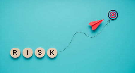A minimalist image with a bright blue background. The word "RISK" is spelled out on four wooden circular blocks. A red paper airplane is shown in mid-flight, following a dashed line path from the blocks towards a bullseye target in the upper right corner of the frame.の写真素材