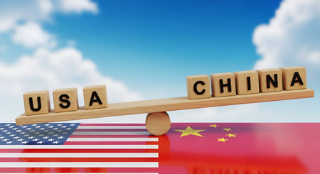 A wooden seesaw is balanced on top of the American and Chinese flags, with wooden blocks spelling "USA" on one side and "CHINA" on the other. The seesaw is tilted, suggesting an imbalance or ongoing negotiation between the two countries. The background is a bright blue sky with clouds.の写真素材