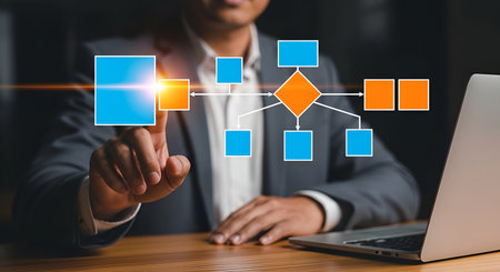A man in a suit is pointing at a digital flowchart displayed on a screen. The flowchart consists of interconnected blue and orange squares and diamonds, illustrating a process or workflow. A laptop is visible on the right. The image conveys business planning, process management, and digital strategy.の写真素材