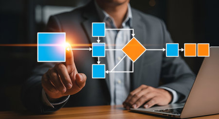 A businessman in a suit is shown from the chest down, his finger pointing towards a glowing blue square on a digital flowchart. The flowchart consists of blue squares and an orange diamond, connected by white lines, extending across the screen. A laptop is visible on the right side of the wooden desk.の写真素材