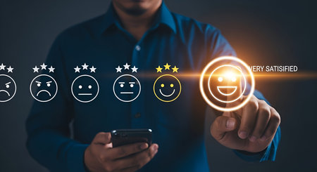 A person wearing a blue shirt holds a smartphone and uses their finger to touch a glowing icon of a happy face with three stars, indicating "VERY SATISFIED". To the left are five emoji faces representing a range of customer satisfaction from angry to happy, with corresponding star ratings. The background is dark and out of focus.の写真素材