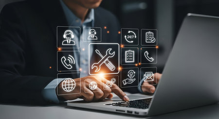 A person in a dark suit jacket is using a laptop. Above the keyboard, a grid of glowing digital icons is displayed, representing various aspects of customer service and support. These include icons for a headset user, a phone, a globe, a wrench and screwdriver, a checklist, a document, a phone receiver, gears, a star with a hand, and an envelope.の写真素材