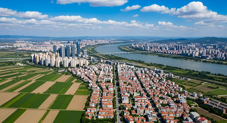 An expansive aerial view shows a modern city with numerous buildings and skyscrapers situated alongside a wide river. A bridge spans the river, connecting different parts of the urban area. In the foreground and to the left, neatly arranged agricultural fields with distinct patterns are visible, contrasting with the urban development. The sky is blue with scattered white clouds.の写真素材