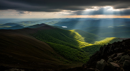 A breathtaking mountain landscape bathed in dramatic light. Sunbeams pierce through dark, heavy clouds, illuminating patches of lush green forests and rolling hills in the valleys below. The contrast between light and shadow creates a moody and atmospheric scene, with a rocky foreground adding texture and depth to the expansive vista.の写真素材