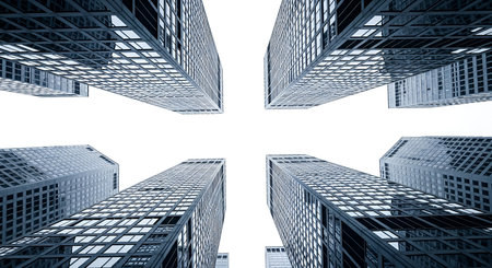 A dramatic low-angle shot looking directly up at a cluster of modern skyscrapers. The buildings are characterized by their glass facades and repetitive geometric window patterns. The perspective emphasizes their height and imposing structure. The sky above is a bright, featureless white, creating a stark contrast with the dark, angular lines of the architecture.の写真素材