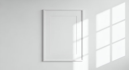 A simple white rectangular picture frame hangs on a plain white wall. Sunlight streams through a window, casting a grid-like shadow pattern across the wall and the frame. The frame is empty, offering a clean space for text or an image.の写真素材