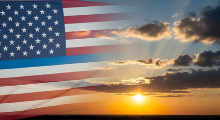 An American flag is superimposed over a dramatic sunset sky. The flag's red and white stripes and blue field with stars are visible, partially transparent against the vibrant orange and yellow hues of the setting sun. Rays of light break through the scattered clouds, illuminating the horizon.の写真素材