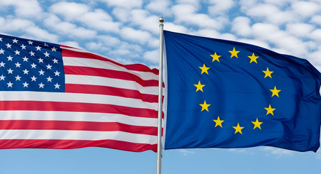 The American flag and the European Union flag are shown waving side-by-side on flagpoles against a blue sky with white clouds. The US flag features its iconic stars and stripes, while the EU flag displays its circle of yellow stars on a blue background. Both flags are captured in motion, suggesting a sense of dynamism.の写真素材