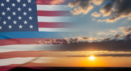 A composite image showing the American flag superimposed over a vibrant sunset sky. The flag's stars and stripes are clearly visible, with its fabric appearing to wave gently. The background features a dramatic sunset with warm orange and yellow hues, illuminated by the setting sun peeking through scattered clouds. The horizon line is visible in the distance.の写真素材