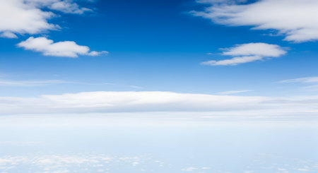 A serene and expansive view of a bright blue sky dotted with scattered, wispy white clouds. The image captures a sense of vastness and openness, as if viewed from a high altitude or during a flight. The lower portion of the image shows a soft, hazy layer of clouds or atmospheric diffusion, creating a gentle gradient of blue.の写真素材