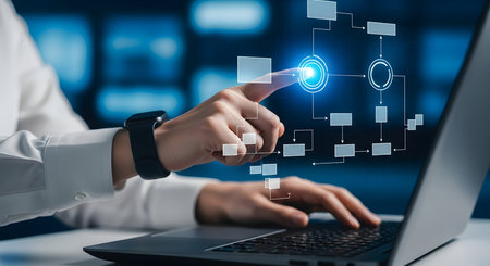 A close-up shot of a person's hand with a smartwatch, touching a glowing blue button on a futuristic digital interface. The interface displays a flowchart with interconnected boxes and lines, suggesting a complex system or process. A laptop is visible in the foreground, with the person's other hand on the keyboard.の写真素材