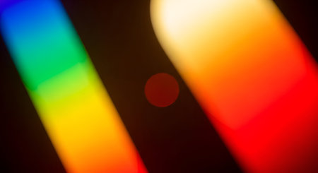 An abstract image featuring blurred bands of vibrant rainbow colors ? red, orange, yellow, green, and blue ? against a dark background. A small, out-of-focus red circle is visible in the center. The colors are soft and luminous, creating a dreamy and artistic effect.の写真素材