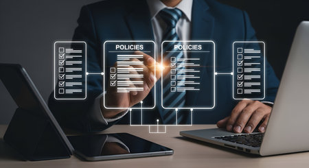 A man in a dark suit and tie is shown at a desk, interacting with a glowing digital interface. He is pointing at screens displaying lists of "POLICIES" with checkboxes, suggesting a review or approval process. A tablet and a laptop are visible on the desk, indicating a modern business setting focused on digital document management.の写真素材