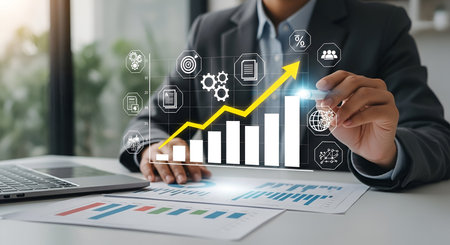 A businessman in a suit jacket and blue shirt is shown at a desk, using a stylus to draw an upward yellow arrow on a holographic bar graph. The graph represents financial growth and is surrounded by digital icons representing business processes and data. A laptop and financial reports are on the desk.の写真素材